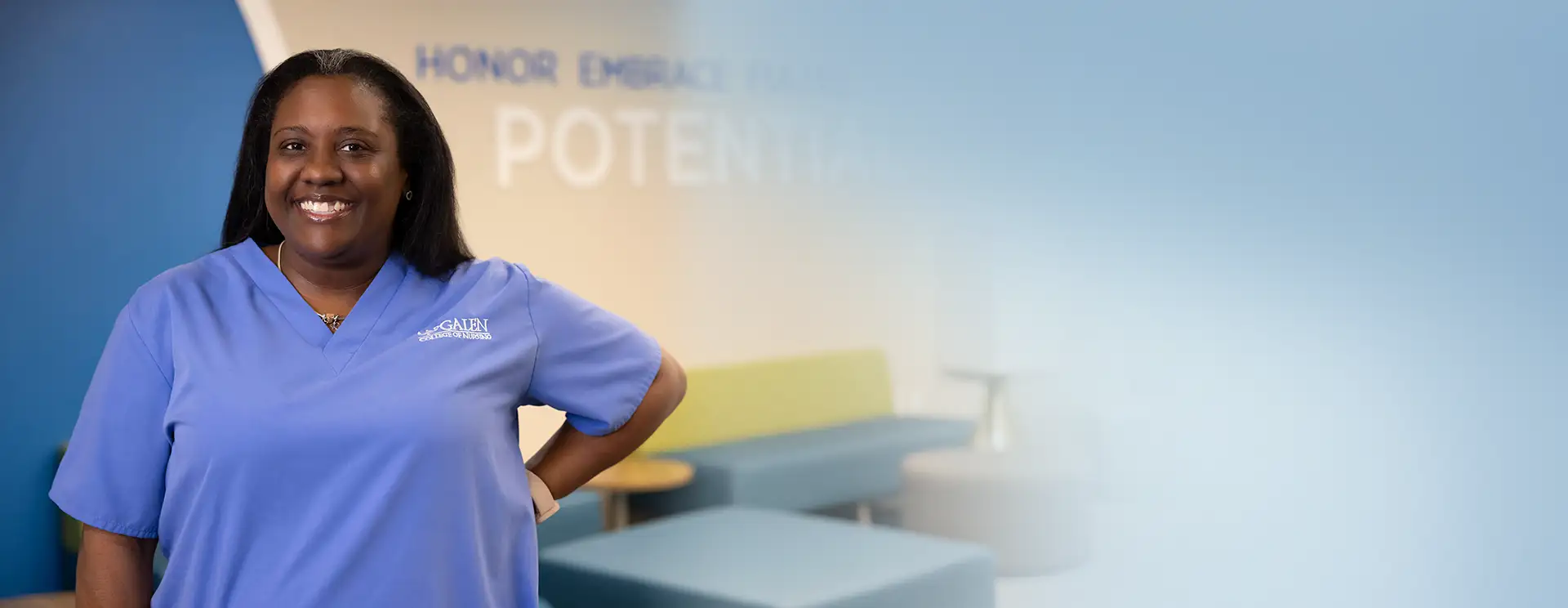 A smiling woman with dark hair, wearing a light blue scrub top with a 'GALEN COLLEGE OF NURSING' logo, poses with one hand on her hip. She has silver strands in her hair and a necklace. In the blurred background, there's a blue and tan wall with large, light-colored text that reads 'HONOR EMB[RACE] POT[ENTIAL]' and a modern sitting area with a green sofa.