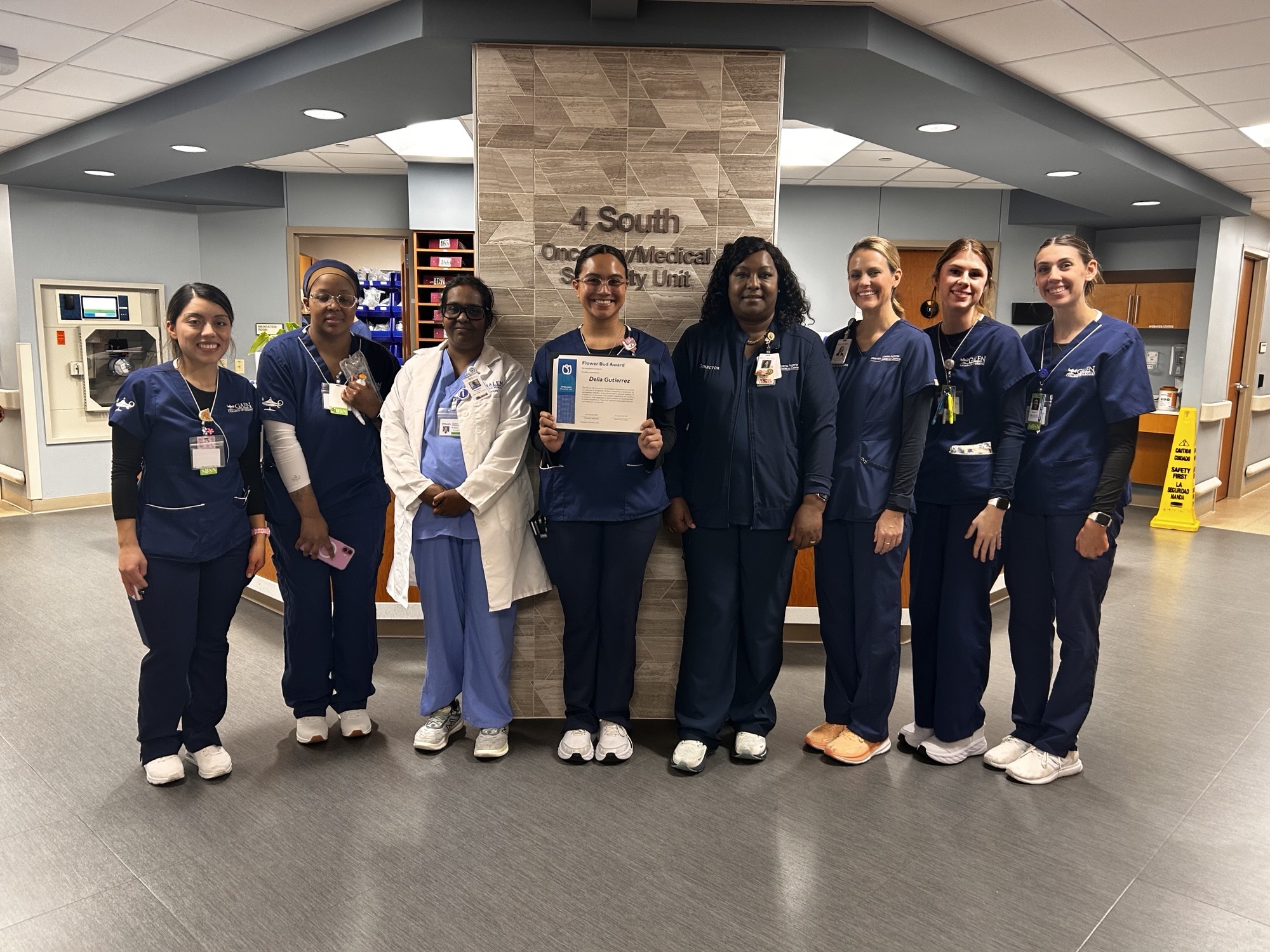 A group of healthcare professionals in navy and white scrubs standing together in a hospital hallway in front of a sign that reads “4 South Oncology Medical Specialty Unit.” One person in the center is holding a certificate.