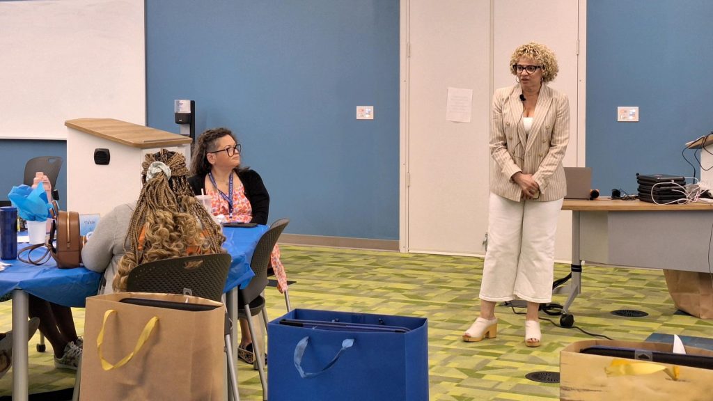 A woman faces the room which has people sitting at tables. She appears to be the main focus of the attendees. 