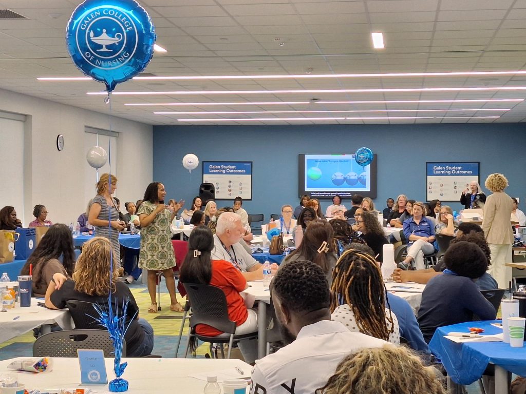 A large bright room with people sitting at tables facing the panelists. A few people are standing and people look engaged with the speakers. 