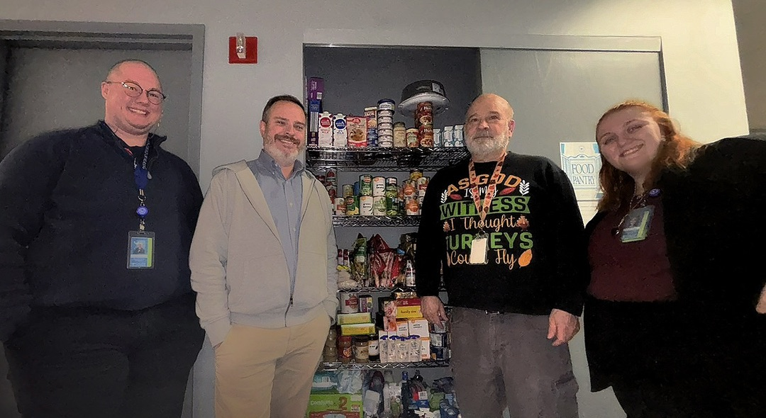Four people standing in front of a stocked food pantry shelf filled with canned goods, boxed items, and other non-perishable food.