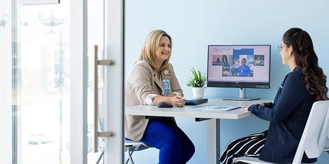 Two women sit across from each other at a desk in an office. One woman, smiling, is wearing a light brown cardigan and blue pants, and the other is wearing a black blazer and striped pants, looking at a computer monitor displaying multiple images of people.