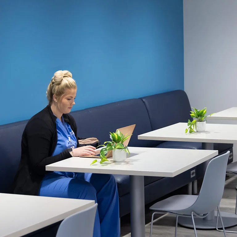 A female nursing student with blonde hair pulled back in a bun, wearing blue scrubs and a black cardigan, sits on a dark blue bench at a white table. She is focused on typing on a laptop. Small potted plants decorate the tables, and a bright blue wall is in the background.