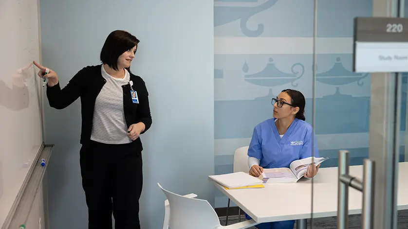 An instructor writing on a white board in a study room, while a student in scrubs watches her