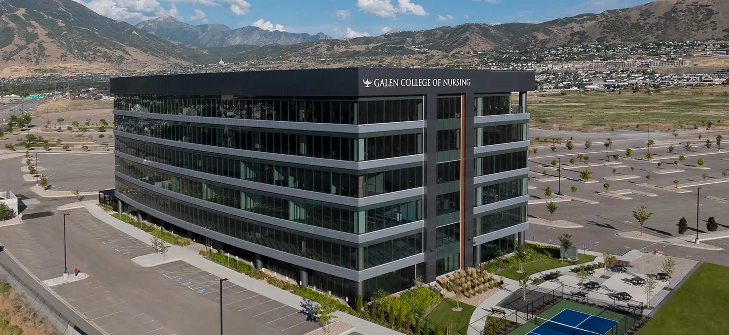 An aerial view of a large, modern Galen College of Nursing building in Salt Lake City, UT with a black and gray facade, extensive glass windows, and a black roof. It's surrounded by a large parking lot, some green landscaping, and mountains in the background. A tennis court is visible on the right.