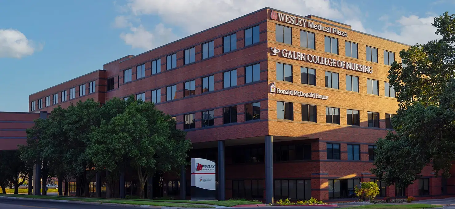 An exterior view of a large, multi-story brick building that houses 'WESLEY Medical Plaza', 'GALEN COLLEGE OF NURSING', and 'Ronald McDonald House'. The building has numerous dark windows and is surrounded by green trees and a paved area. A sign for 'WESLEY Medical Plaza' is visible in the foreground on the left.