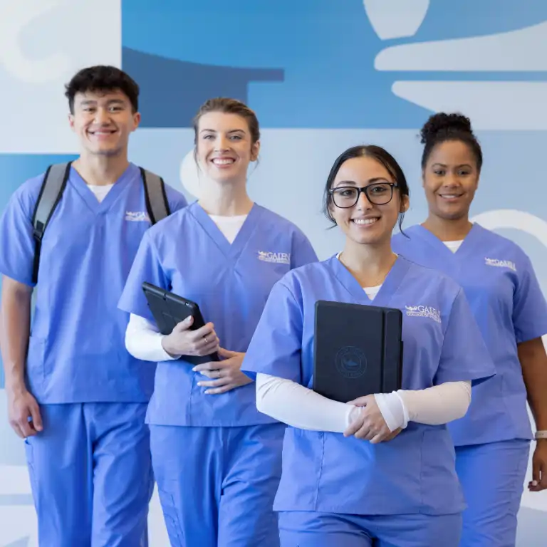 Four smiling nursing students in blue scrubs pose in a brightly lit hallway. Three women and one man, all wearing 'GALEN' branded scrubs, are holding notebooks or tablets. The background is a light blue and white wall with abstract shapes.