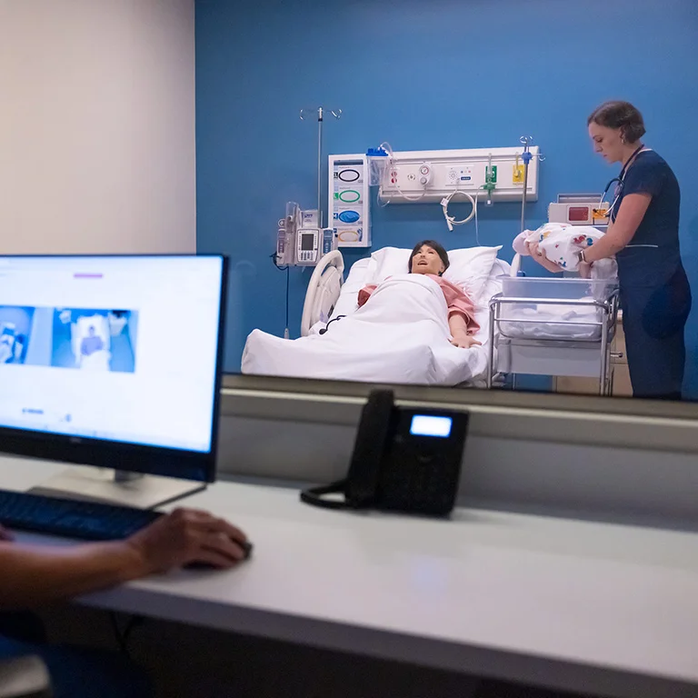A wide shot of a nursing simulation lab with a student in blue scrubs tending to a baby manikin in a bassinet. Another patient manikin lies in a bed behind her. In the foreground, a person's arm is visible using a computer mouse at a desk with a monitor displaying medical images and a phone.