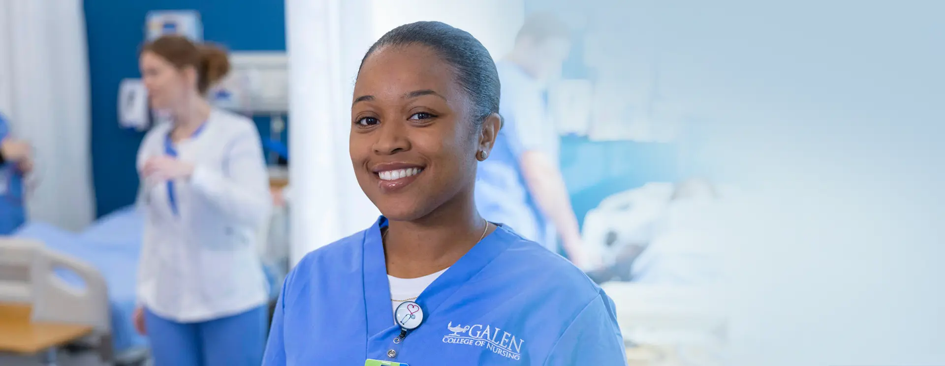 Female nursing student smiling at camera with simulation lab activities happening behind her.