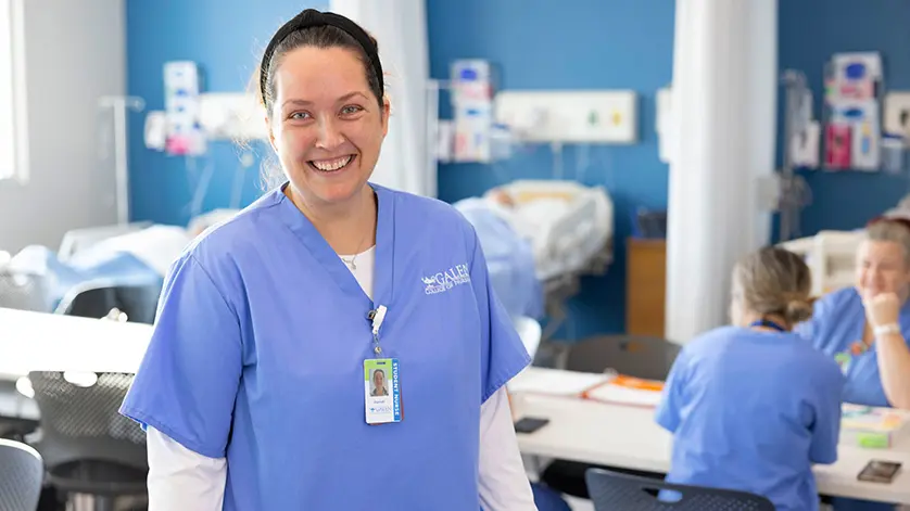Female nursing student smiling at camera with simulation lab activities happening behind her.