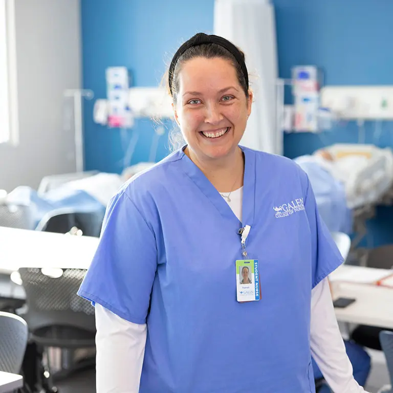 Female nursing student smiling at camera with simulation lab activities happening behind her.