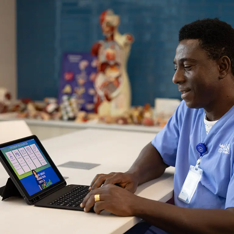 A male Galen student in scrubs looks up from his laptop while studying in a classroom with anatomical models in the background
