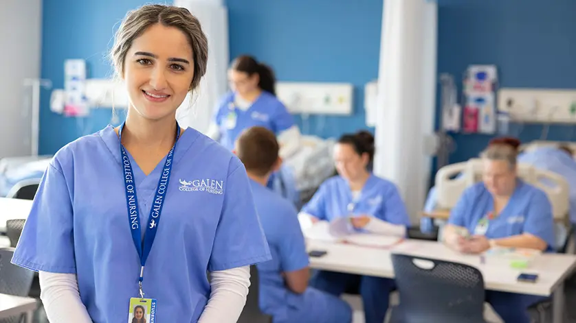 Female nursing student smiling at camera with simulation lab activities happening behind her.