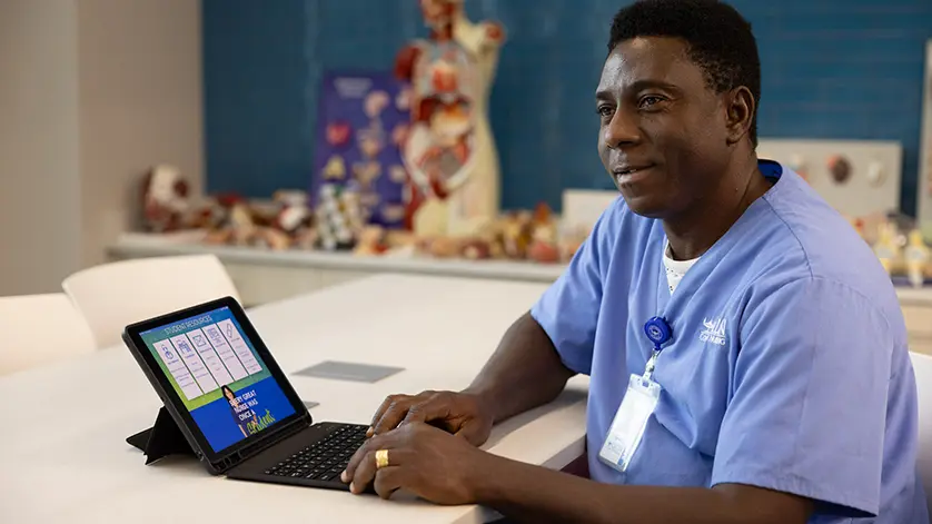 A male Galen student in scrubs looks up from his laptop while studying in a classroom with anatomical models in the background
