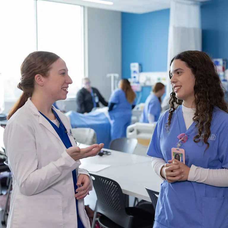 Galen nursing instructor and student in scrubs, talking with each other with simulation lab activities in background