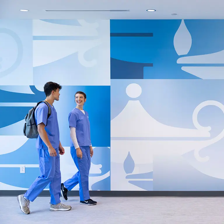 two students, a male and female, walking along a wall at a Galen campus and talking to each other.