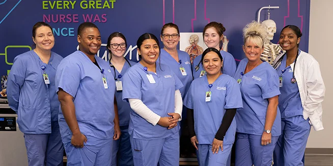 A group of nine diverse nursing students and one instructor, most wearing blue scrubs and ID badges, pose together smiling in front of a purple wall with inspirational text. Anatomical models, including a skeleton and torso, are visible in the background.